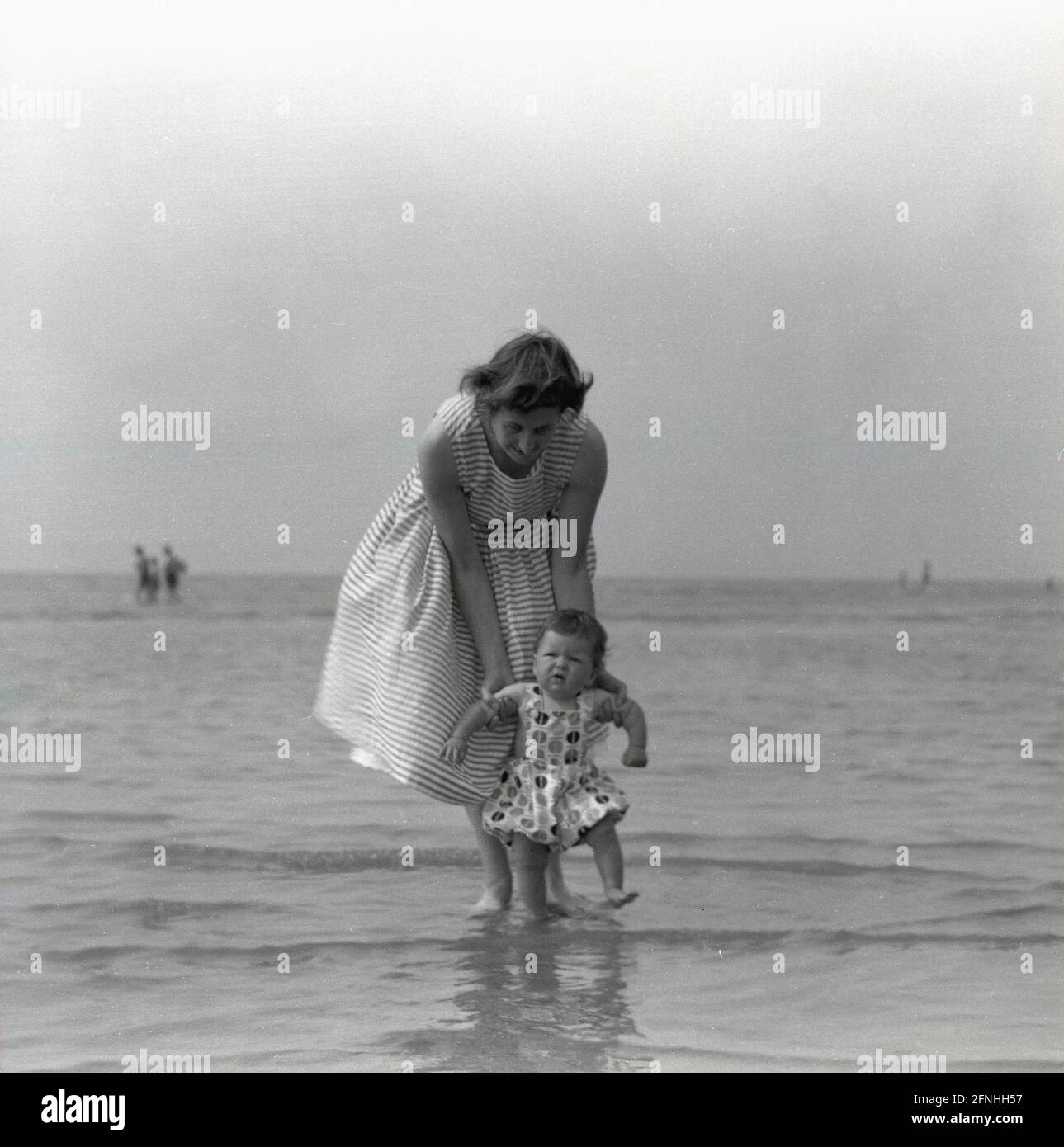 1959, historique, à l'extérieur dans la zone peu profonde de la mer, une mère dans une longue robe de l'époque, tenant son enfant en bas âge comme il a sa première expérience d'une paddle dans l'eau de mer, Angleterre, Royaume-Uni. Banque D'Images