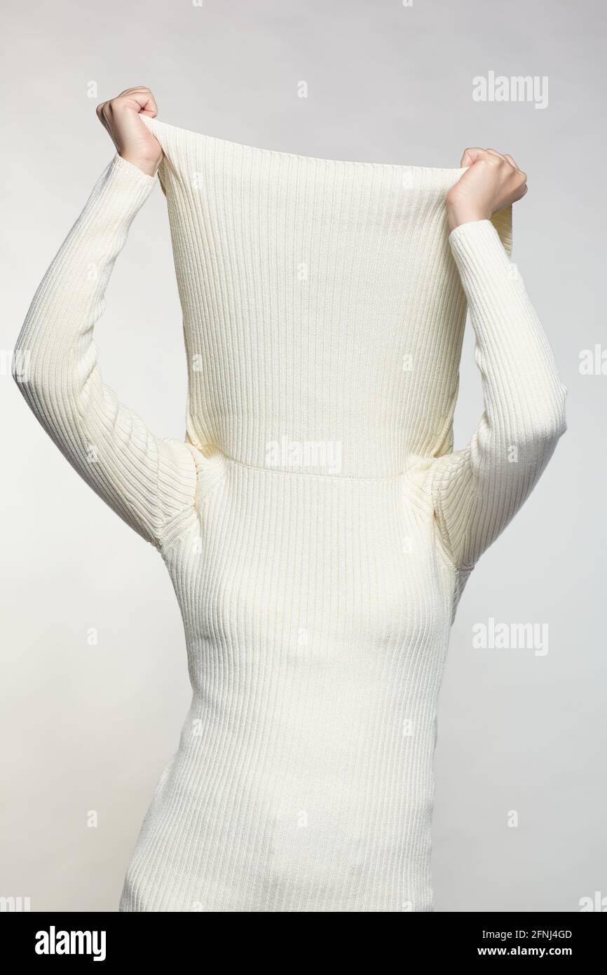 Femme vêtue d'un grand pull blanc en laine sur fond gris. Femme avec les mains sur le col du chandail cache le visage. Banque D'Images