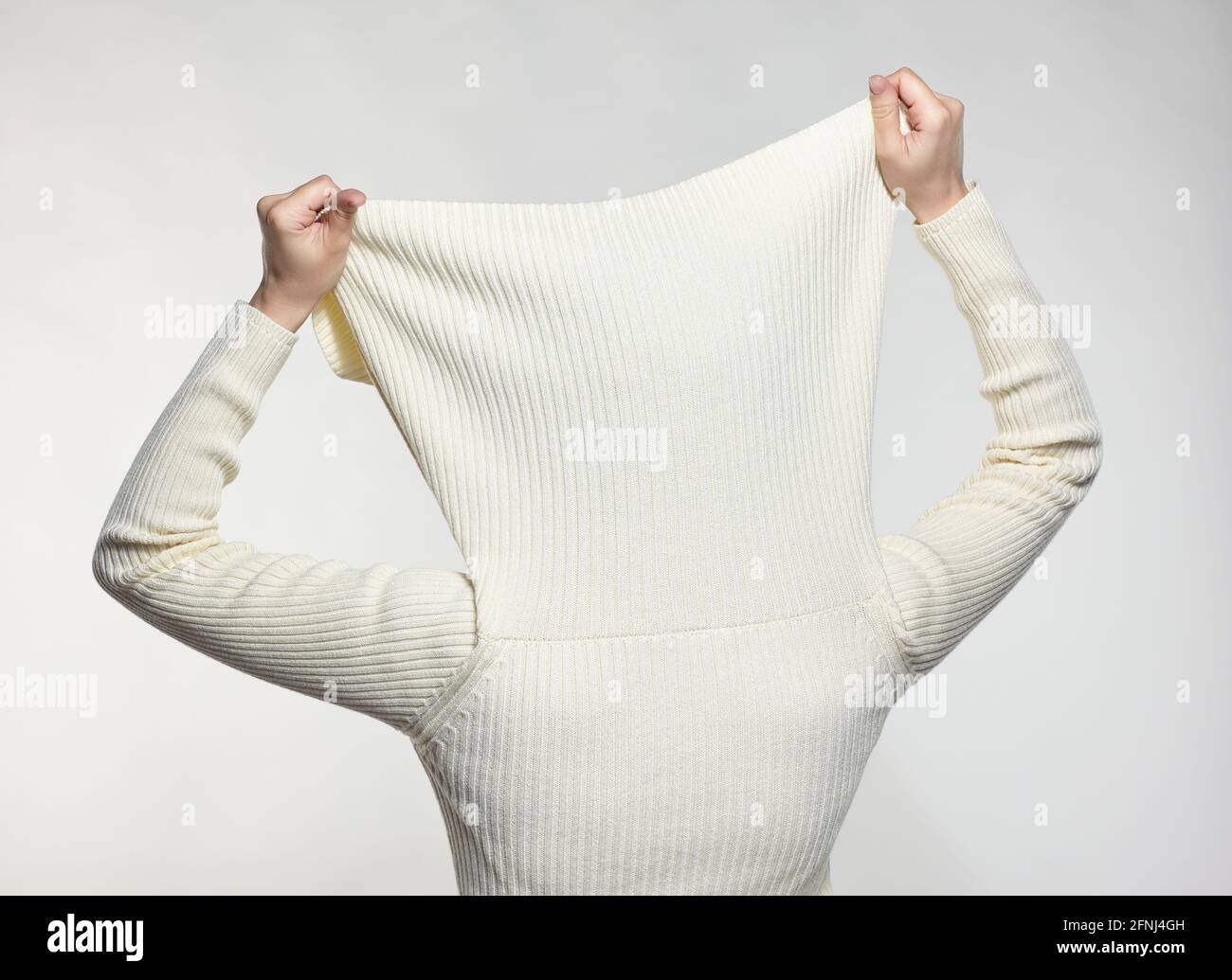 Femme vêtue d'un grand pull blanc en laine sur fond gris. Femme avec les mains sur le col du chandail cache le visage. Banque D'Images