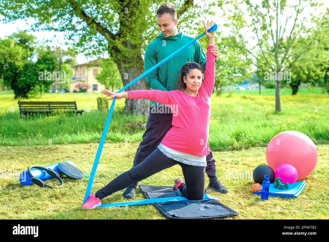 Jeune femme enceinte s'entraîne bras avec résistance élastique bande corde extensible assis sur un ballon de gym. Entraîneur personnel train une fille à l'extérieur dans le parc. Banque D'Images