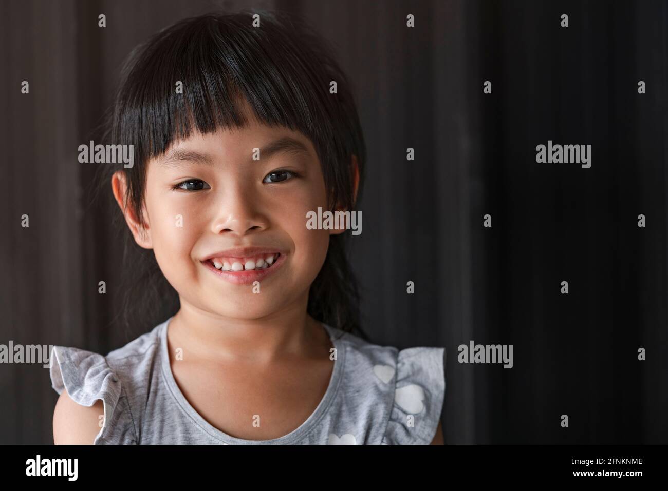 Portrait charmante petite fille asiatique, visage souriant, cheveux noir éclatant, montrant des dents et des gencives blanches saines, fond en métal noir. Espace pour le texte. Banque D'Images