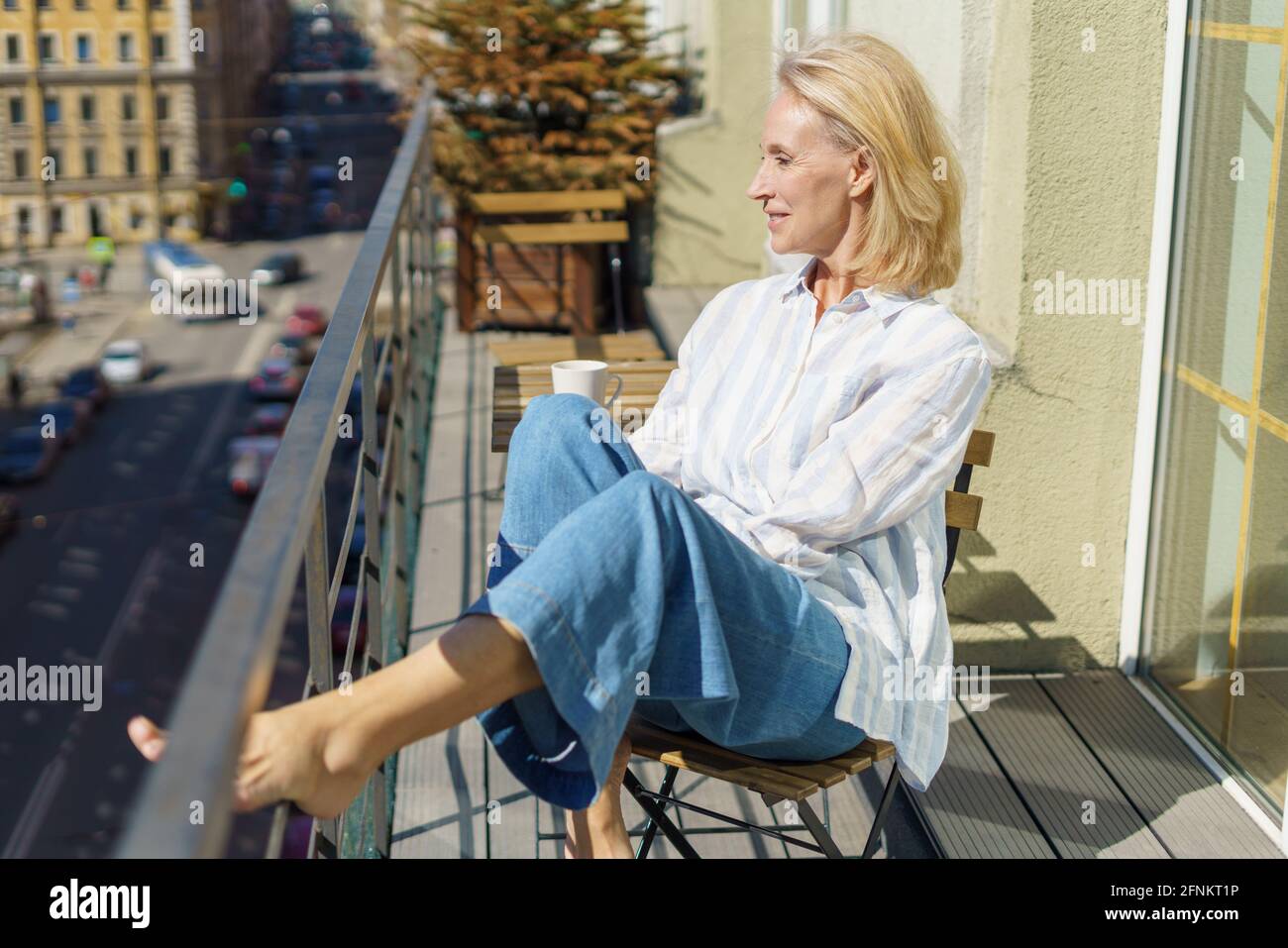 Le matin. Belle femme adulte rêveuse des années 60 dans une tenue décontractée se détendre sur le balcon à la maison, commencer la journée avec le café. Bonne femme âgée assise sur la terrasse et regardant la rue ensoleillée Banque D'Images