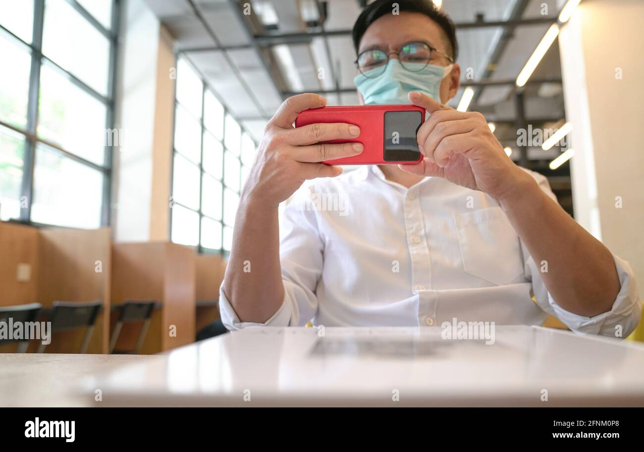 Homme portant un masque facial utilisant son téléphone pour regarder ou prendre une vidéo. Intérieur. Banque D'Images