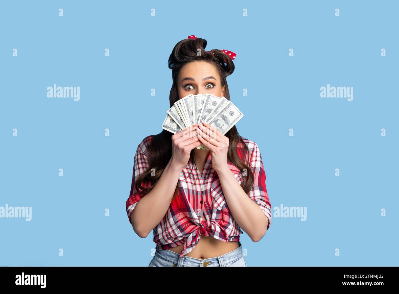 Jeune femme attirante pinup excitée de gagner une grosse somme d'argent, détenant des billets de dollars sur fond bleu studio Banque D'Images