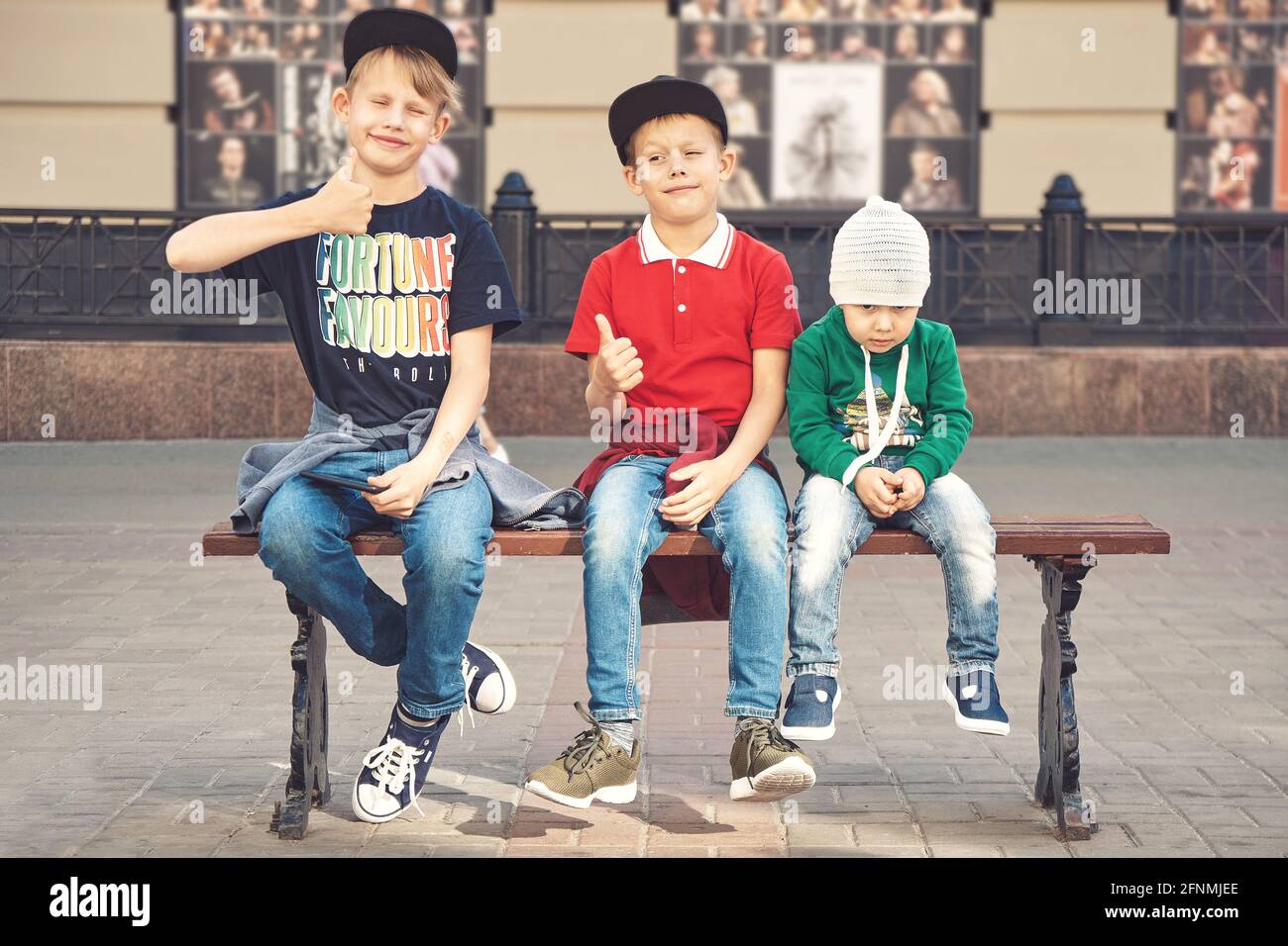 Les frères aînés positifs en casquettes stylées montrent les pouces assis  près petit garçon fatigué sur le banc dans une rue de ville contemporaine  Photo Stock - Alamy