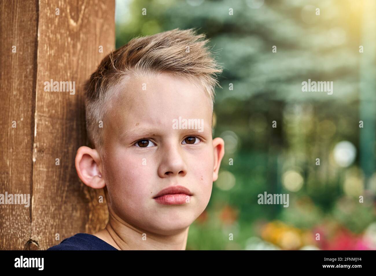 Joli garçon blond avec une coiffure courte s'appuie sur une clôture en bois poser pour caméra dans vert ensoleillé été ville parc extrême gros plan Banque D'Images