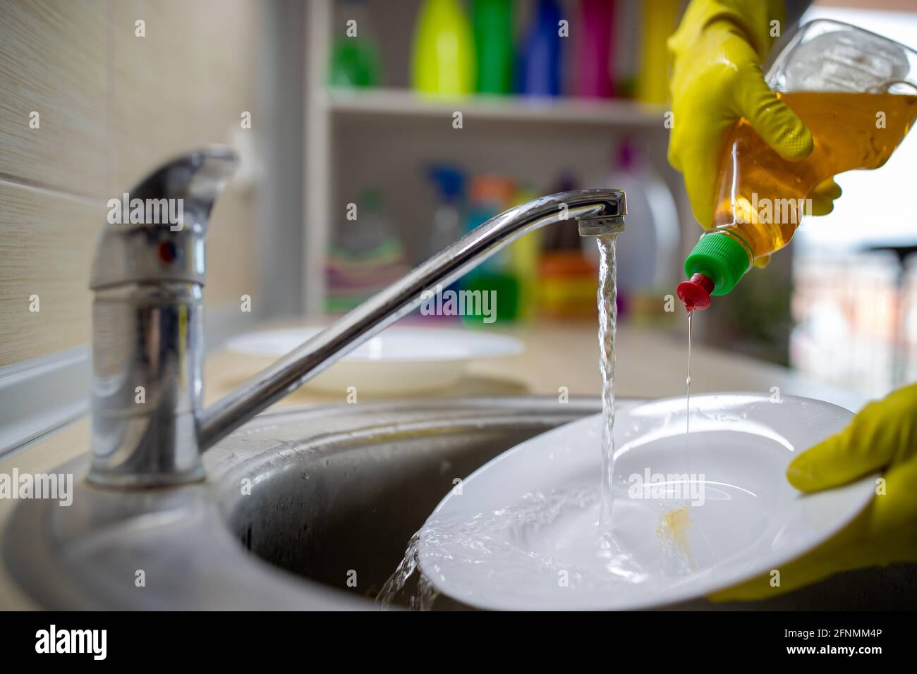 Gros plan des mains des femmes avec des gants versant du détergent liquide sur la plaque pour laver la vaisselle sous l'eau du robinet Banque D'Images