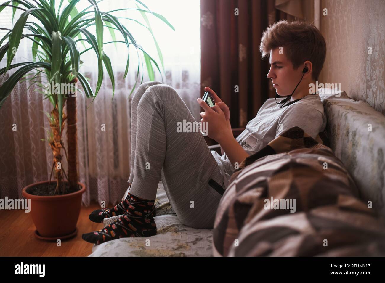 Un jeune garçon, un adolescent, s'assoit sur le canapé à la maison dans un casque et écrit des sms sur un réseau social ou écouter de la musique sur un smartphone. Banque D'Images