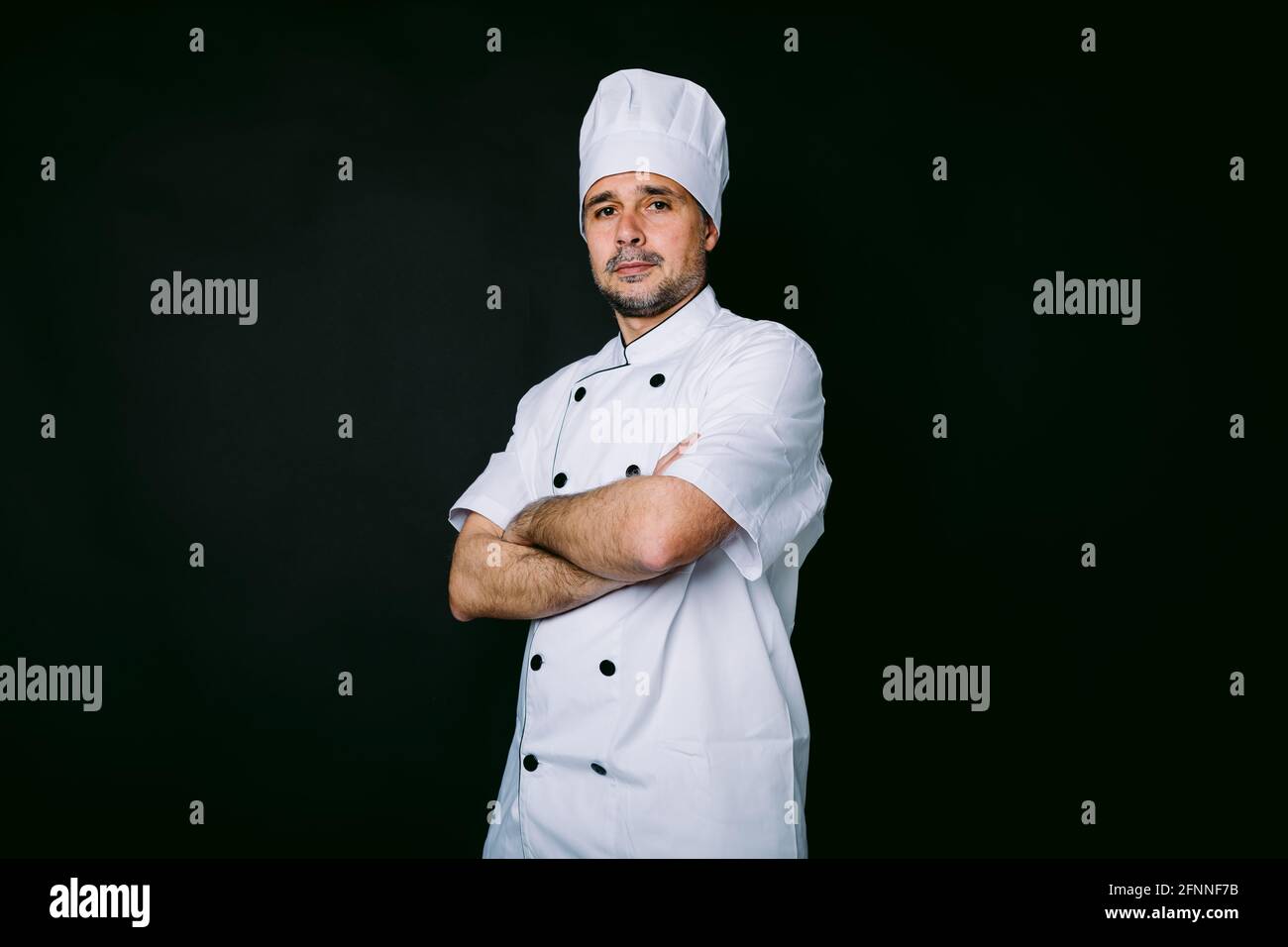 Chef cuisinier portant une veste et un chapeau de cuisine, pose avec des  bras croisés sur fond noir Photo Stock - Alamy