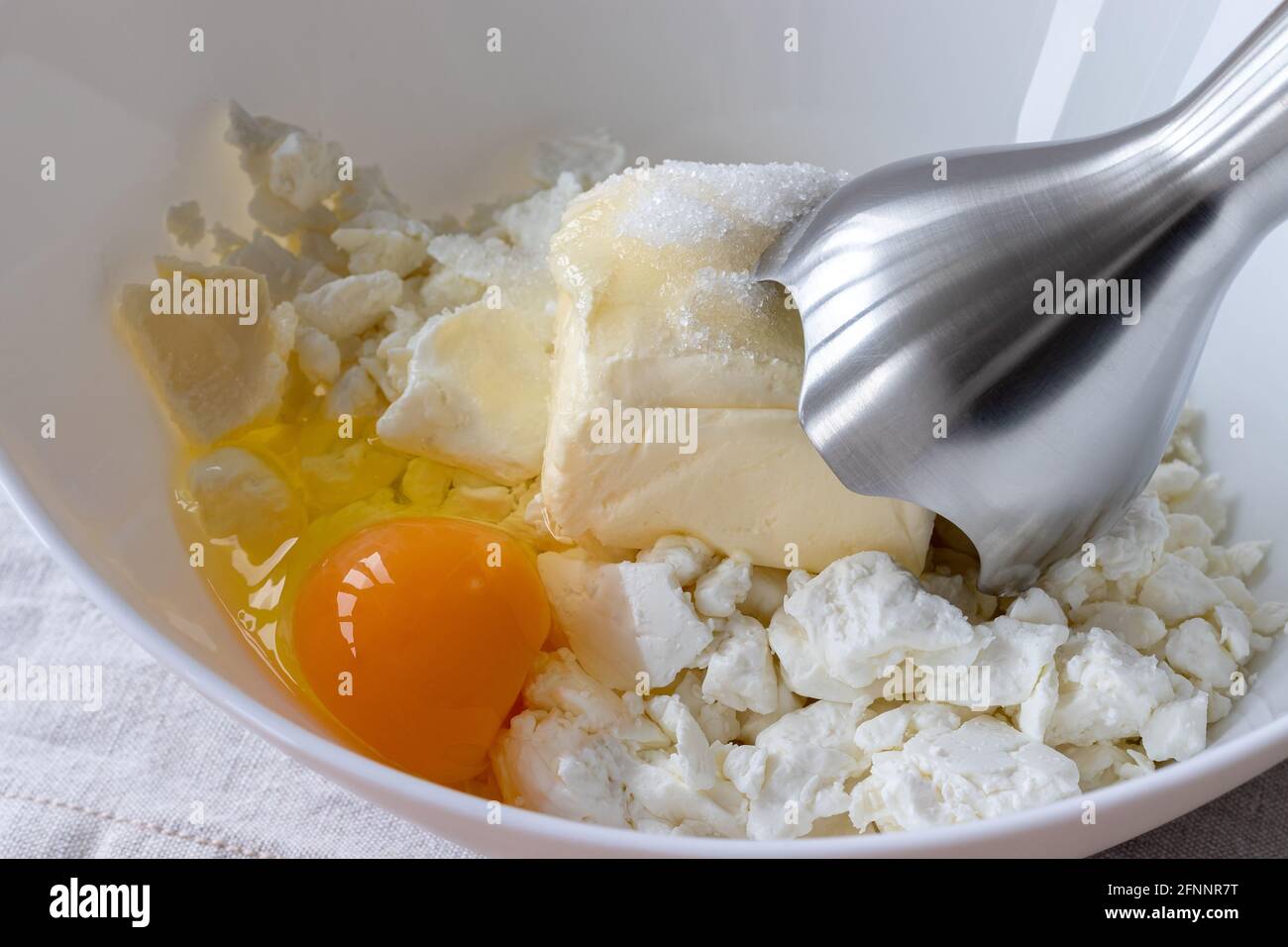 Fromage cottage, beurre, œuf et sucre dans un bol blanc - ingrédients à mélanger avec un mixeur plongeant. Le processus de fabrication de petits pains ou de croissants caillé Banque D'Images