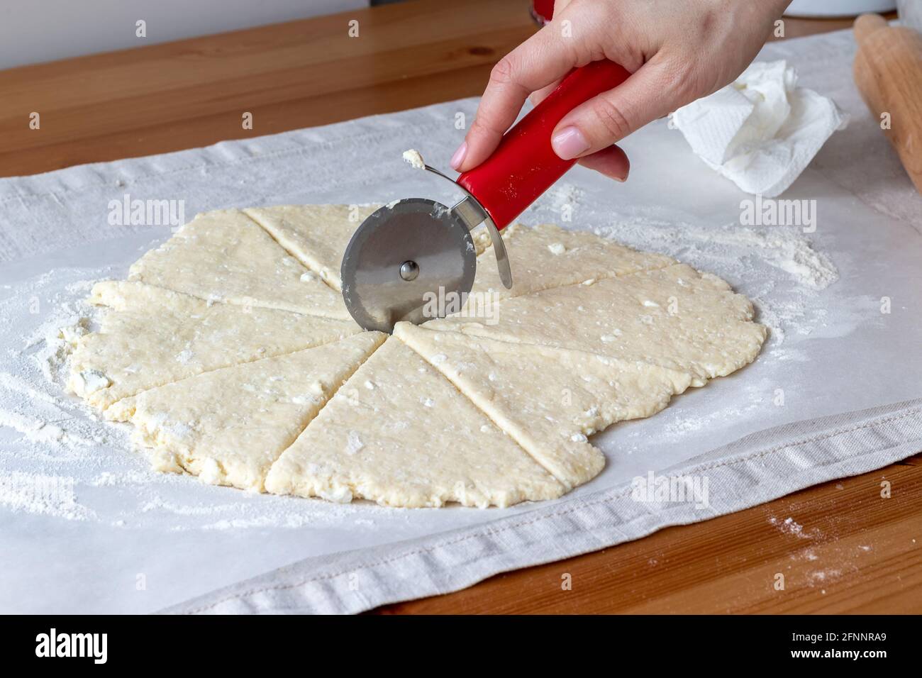 Coupe à la main la pâte à caillé brute roulée avec un couteau à pizza rond en huit triangles sur du papier de cuisson sur une nappe en lin sur une table en bois. Le processus de Banque D'Images