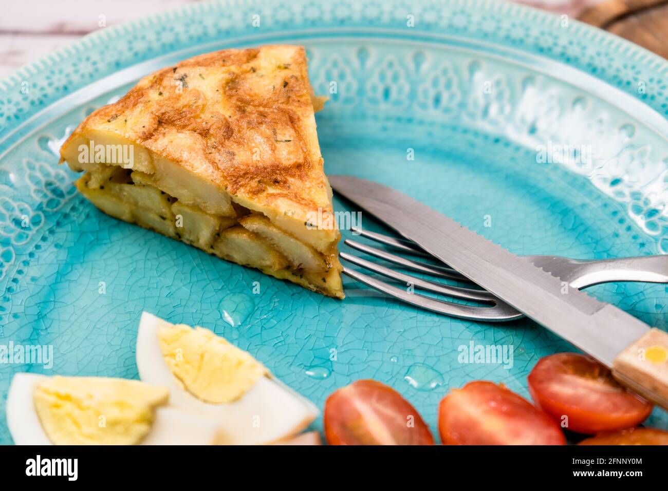 Portion d'une pomme de terre ou omelette espagnole typique sur une assiette bleu clair avec un œuf et des tomates cerises dans un cadre rustique. Espagnol traditionnel Banque D'Images