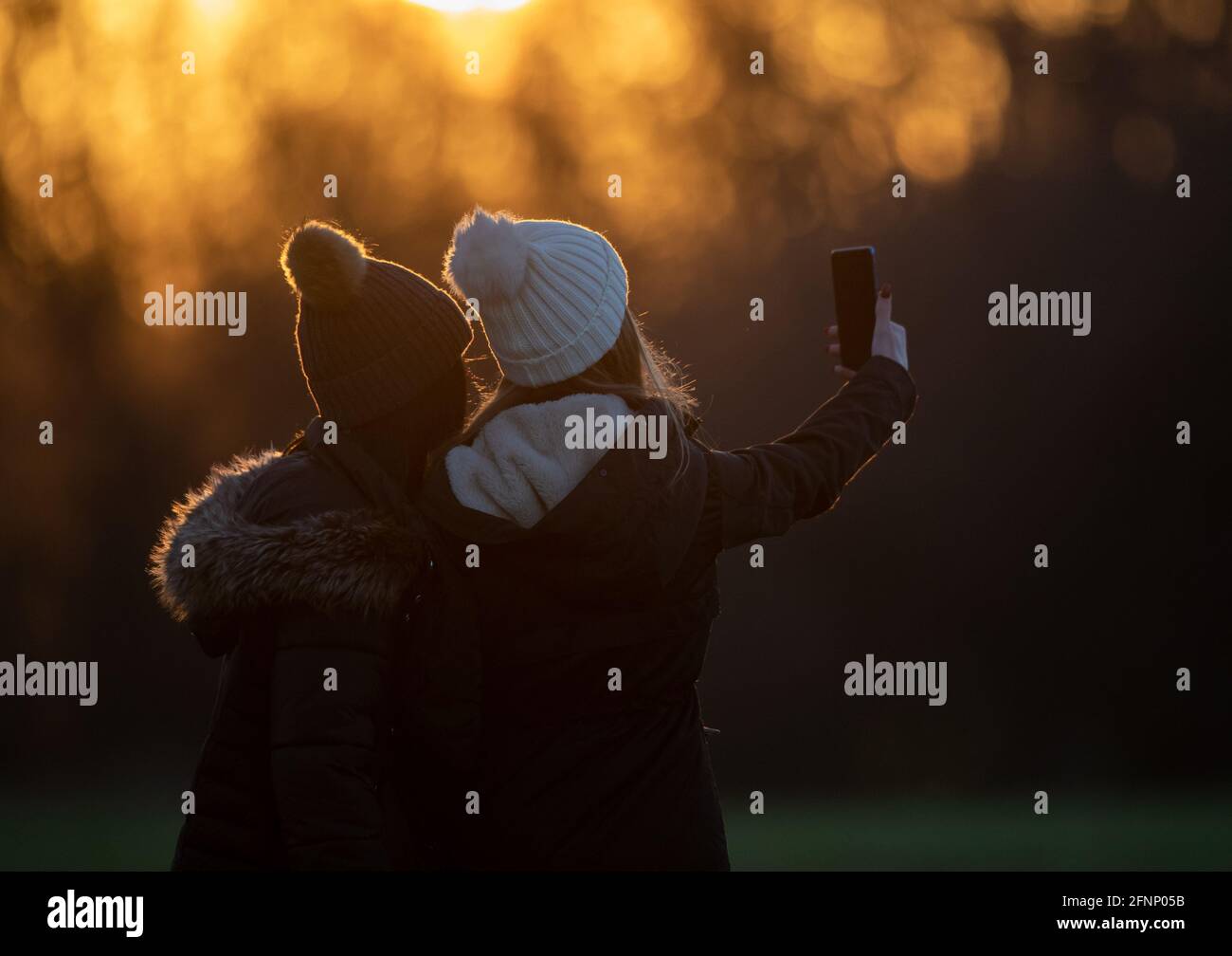 Vue arrière de deux adolescentes en vestes d'hiver et avec des casquettes qui prennent le selfie dans le parc au coucher du soleil Banque D'Images