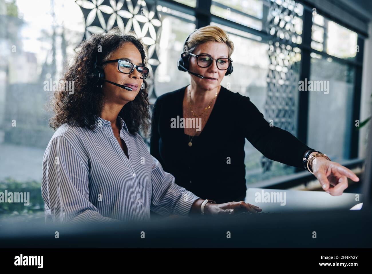 Deux femmes d'affaires portant un casque travaillant ensemble sur un ordinateur. Le responsable pointe vers l'écran de l'ordinateur et discute de quelque chose avec un collègue travaillant chez Banque D'Images