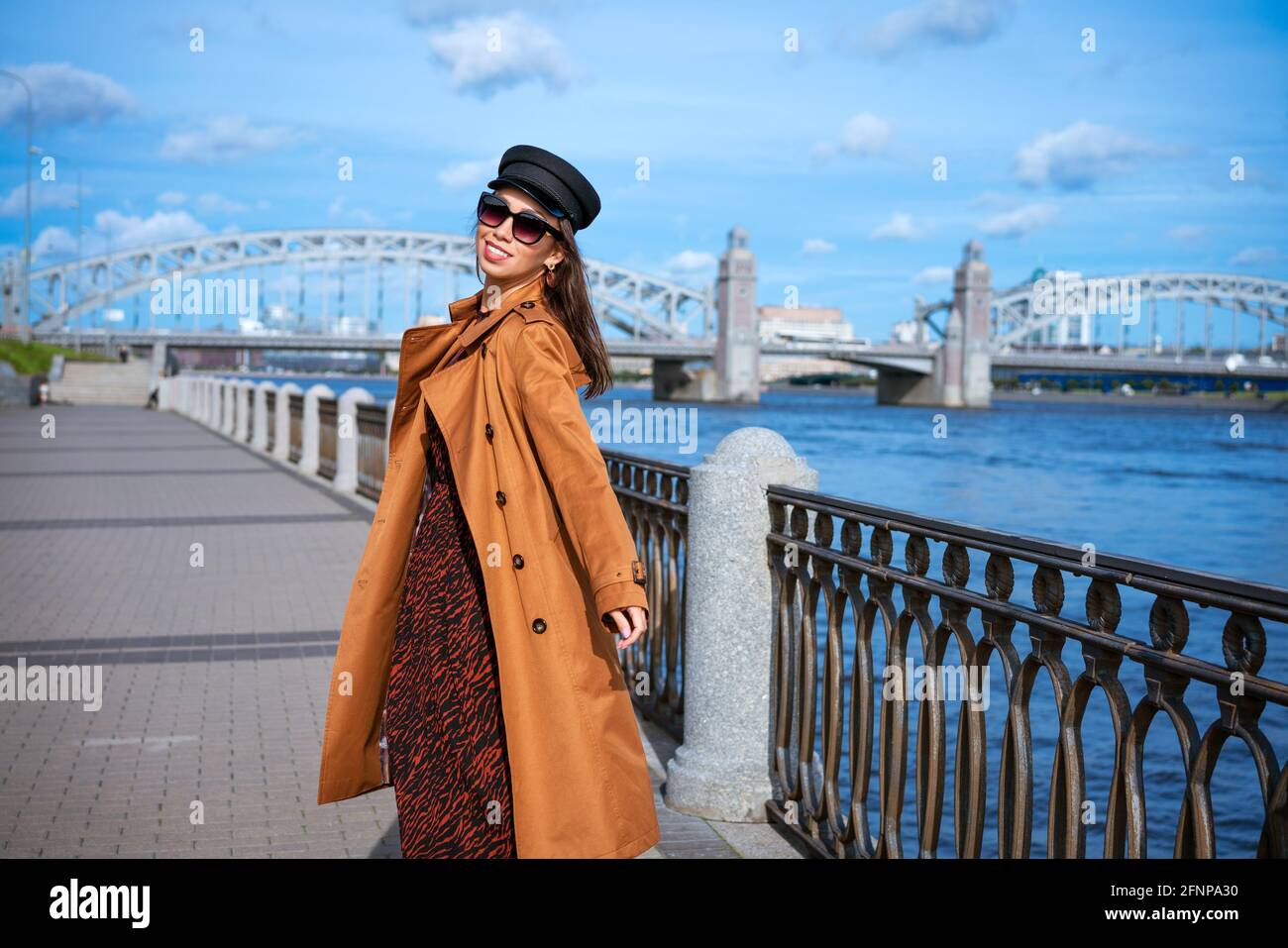 Une jeune femme heureuse d'origine caucasienne, revêtue d'un manteau brun  clair et d'une casquette noire, qui pose sur le remblai sur fond de pont et  de ciel bleu Photo Stock - Alamy