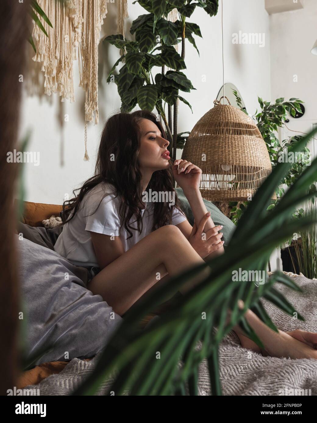 La jeune femme attrayante se détend sur un lit confortable dans une chambre confortable. Style intérieur boho chic avec plantes d'intérieur et macrame Banque D'Images