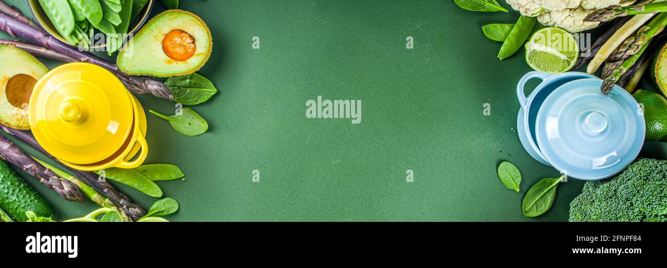Divers ingrédients biologiques de légumes de printemps verts avec des marmites colorées vides sur table de cuisine vert foncé, vue du dessus. Une soupe saine à l'alimentation Banque D'Images