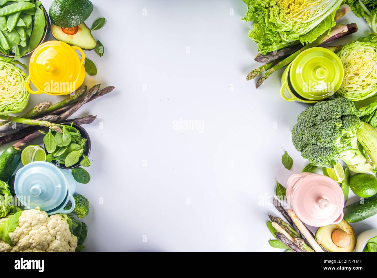 Divers ingrédients biologiques de légumes de printemps verts avec pots de cuisson colorés vides sur table de cuisine blanche, vue du dessus. Soupe saine alimentation, ste Banque D'Images
