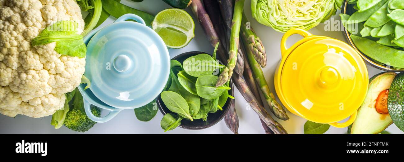 Divers ingrédients biologiques de légumes de printemps verts avec pots de cuisson colorés vides sur table de cuisine blanche, vue du dessus. Soupe saine alimentation, ste Banque D'Images