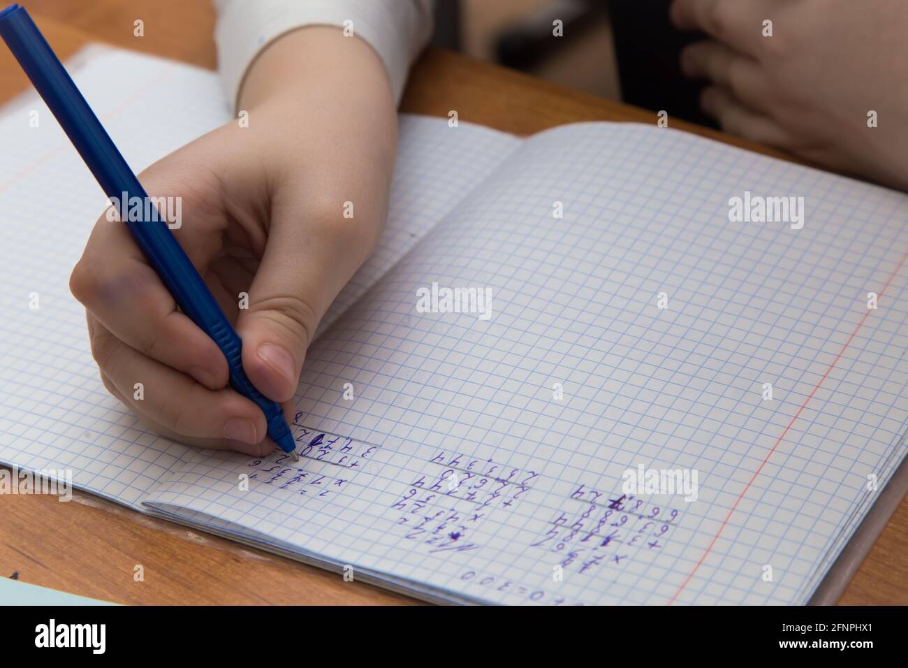 Gros plan de la main d'une fille avec un stylo à bille écrit des chiffres dans un bloc-notes. L'étudiant termine la tâche. Un étudiant résout des exemples en mathématiques à l'école à un bureau. Lumière du jour douce et chaude. Banque D'Images