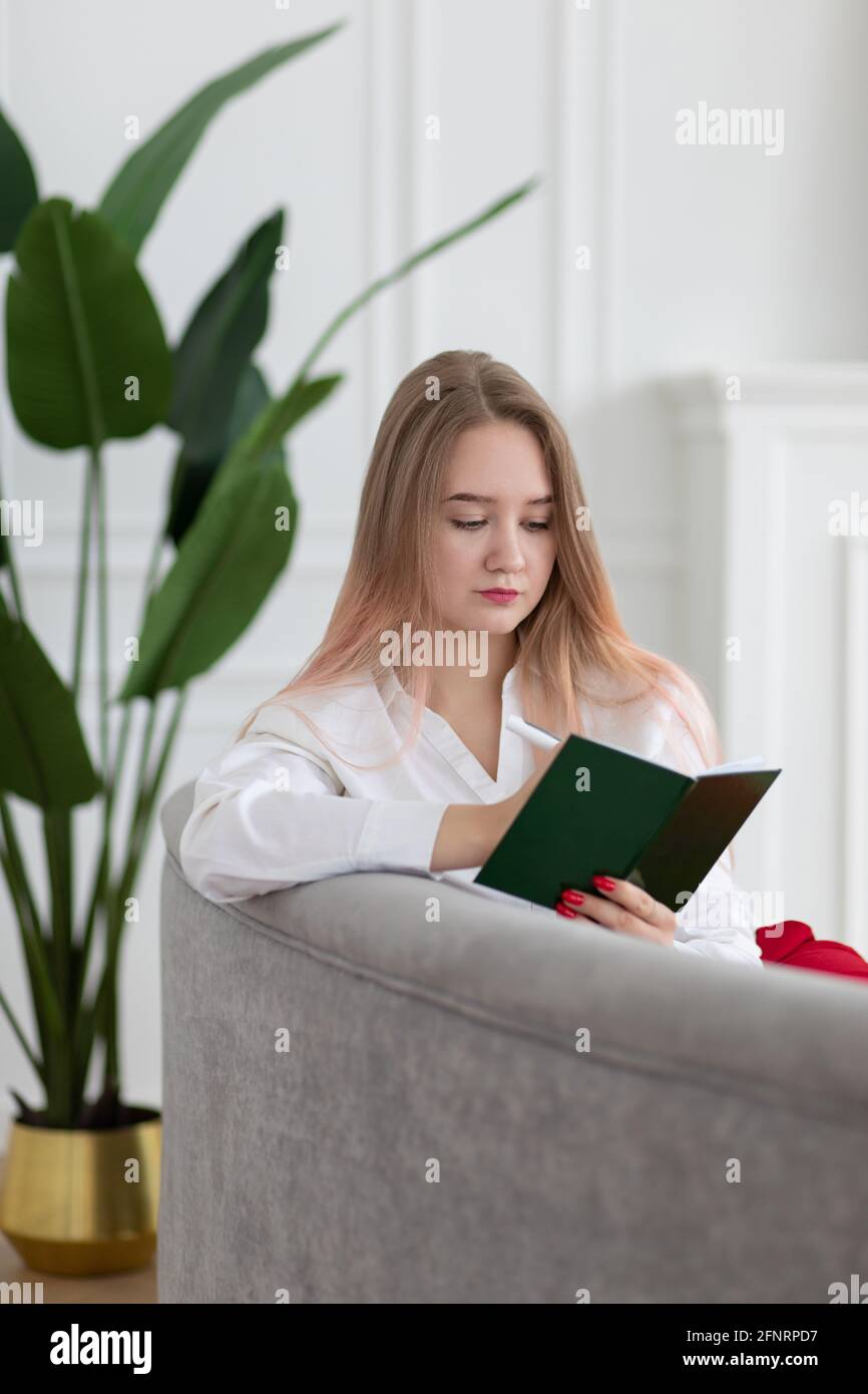 Blonde longue cheveux jeune femme d'affaires caucasienne écrivant dans le bloc-notes de planificateur, journal, planification de travail et d'affaires programme, assis sur un canapé, sho vertical Banque D'Images