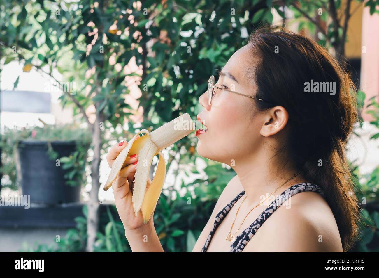 Femme asiatique en verres avec vernis à ongles rouge et un bâton de lèvre manger une banane mûre Banque D'Images Femme asiatique en verres avec vernis à ongles rouge et un bâton de lèvre manger une banane mûre Banque D'Images