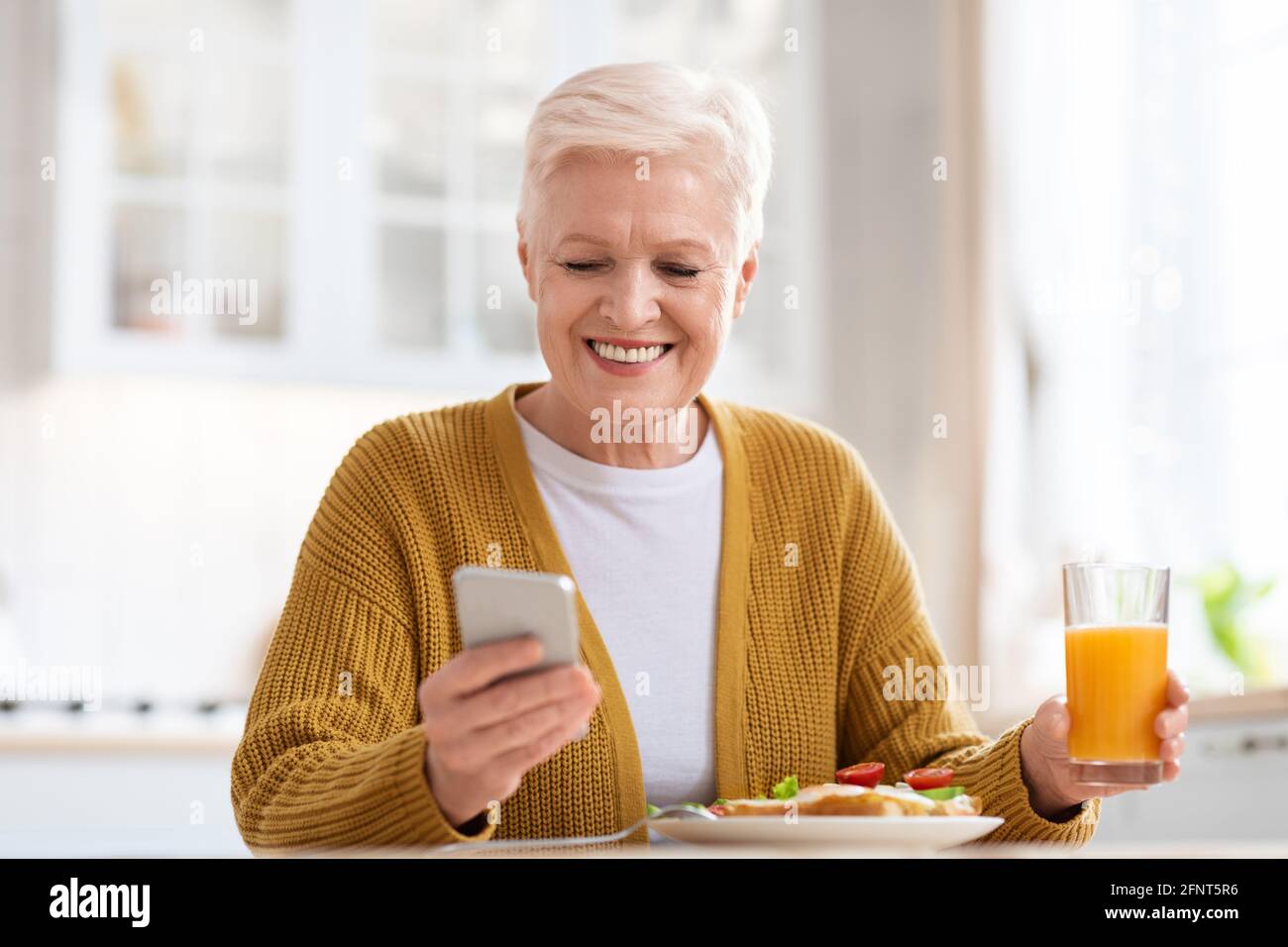 Femme âgée gaie utilisant le téléphone portable tout en déjeuner, gros plan Banque D'Images