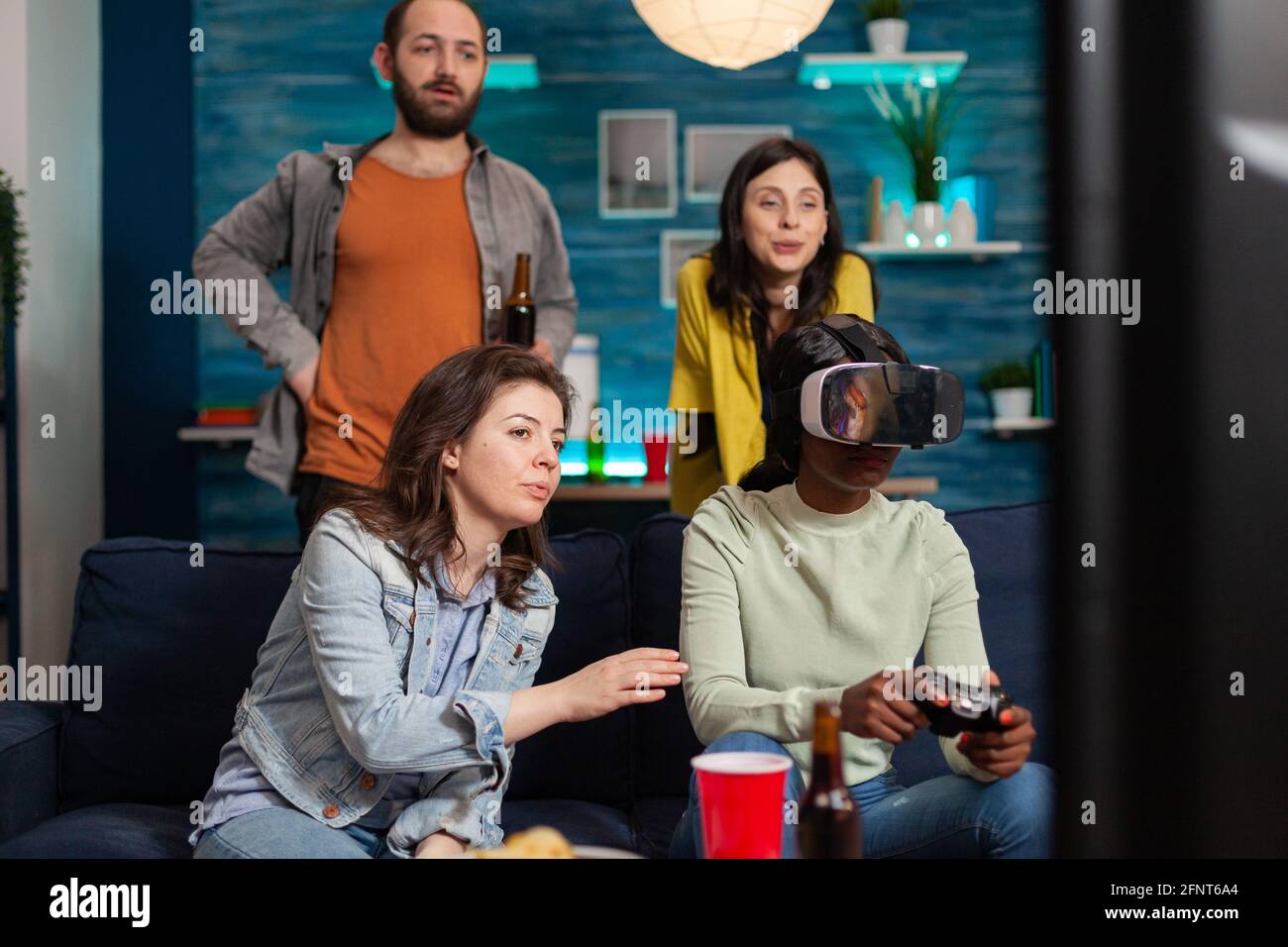 Amis multiethniques qui se socialisent en jouant à des jeux vidéo en faisant l'expérience de la réalité virtuelle à l'aide d'un casque. Groupe de course mixte de personnes qui traînaient ensemble en s'amusant tard dans la nuit dans le salon. Banque D'Images