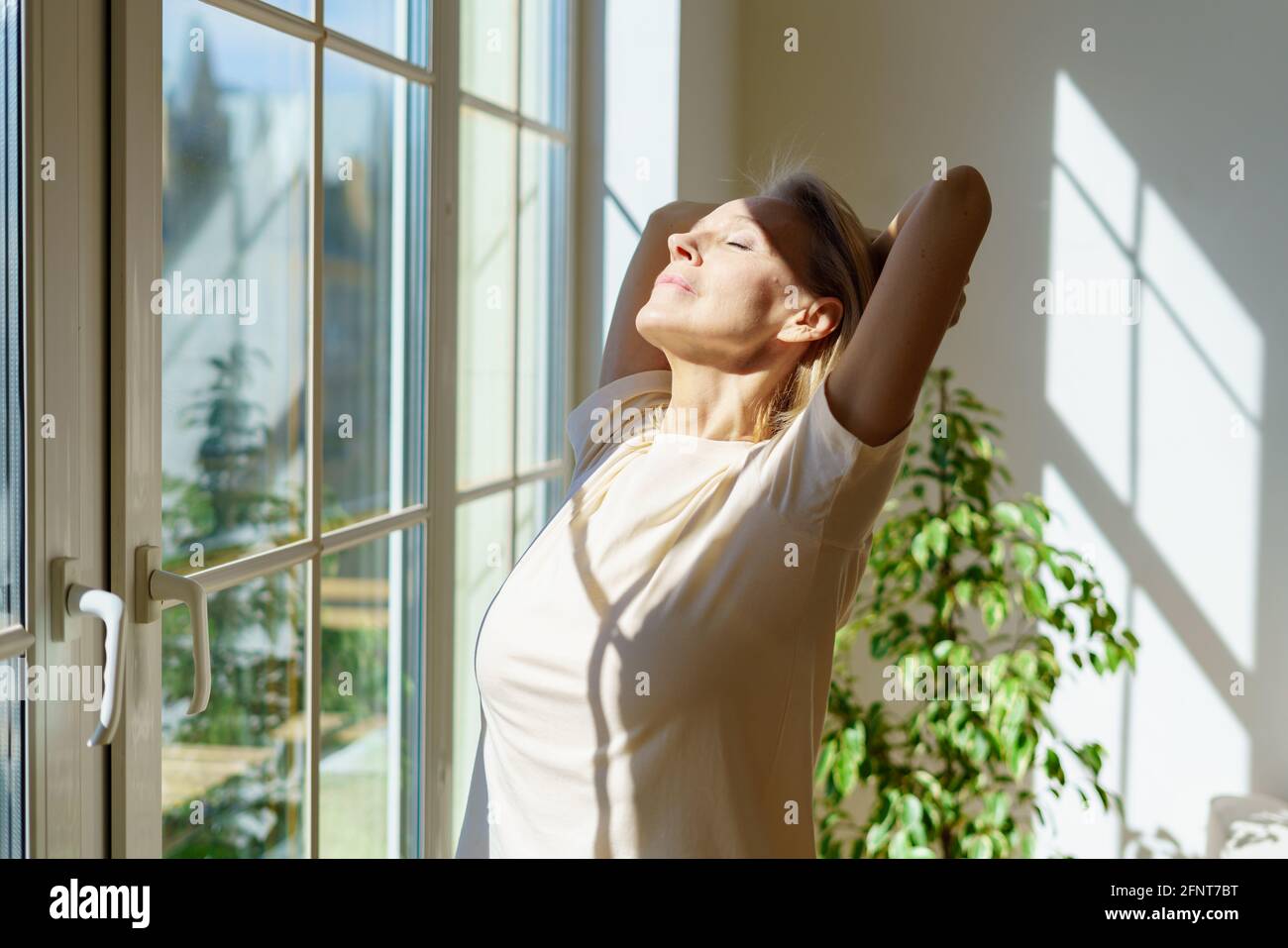 Nouvelle journée. Belle heureuse femme mûre s'étirant le corps et souriant tout en se tenant avec les yeux fermés dans les rayons du soleil qui brillent à travers la fenêtre le matin ensoleillé à la maison. Concept d'optimisme et de bonheur Banque D'Images