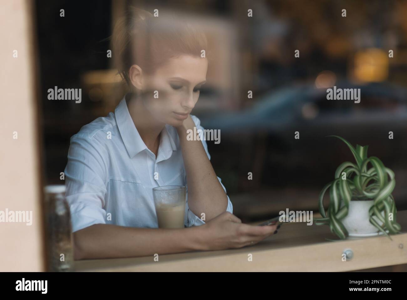 Belle jeune fille s'assoit dans un café près de la fenêtre derrière un verre avec une tasse de café et envoie un message sur le smartphone. Photo à travers verre avec Banque D'Images