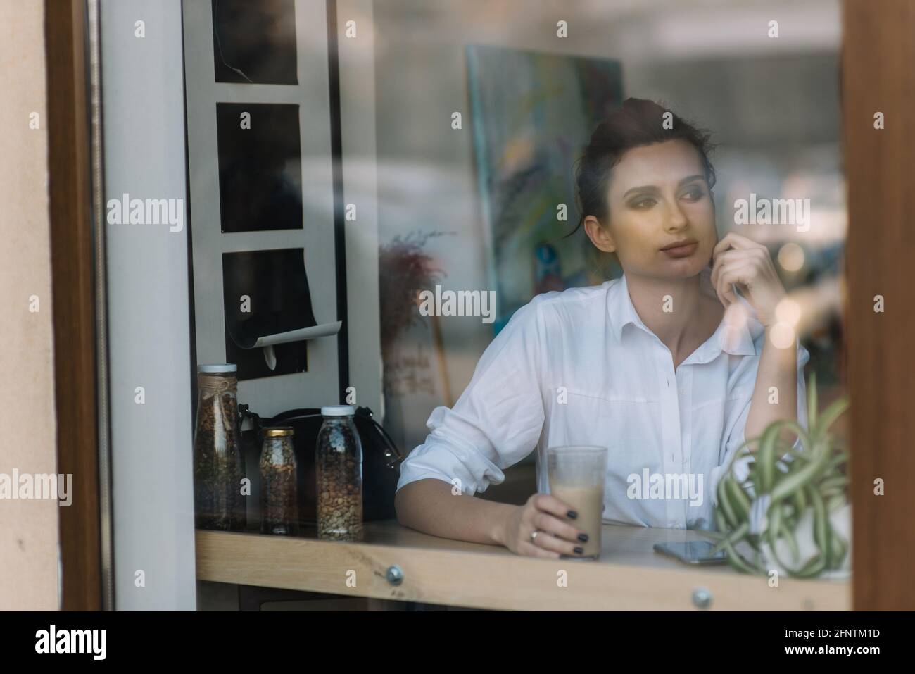 Une belle jeune fille est assise dans un café derrière un verre avec une tasse de café et regarde soigneusement par la fenêtre. Photo à travers le verre avec le refle de la rue Banque D'Images