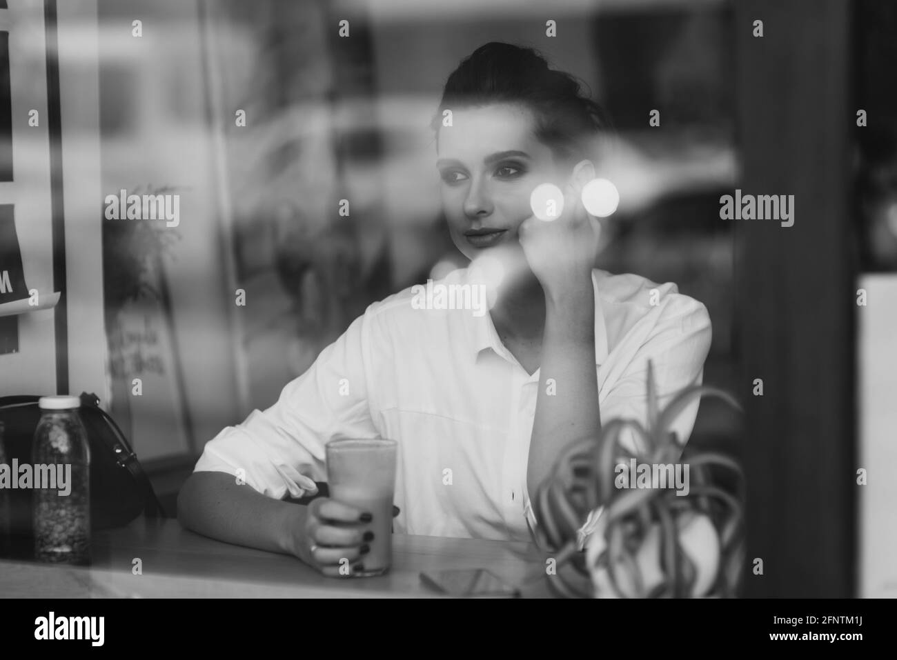 Une belle jeune fille est assise dans un café derrière un verre avec une tasse de café et regarde soigneusement par la fenêtre. Photo d'art noir et blanc via gla Banque D'Images