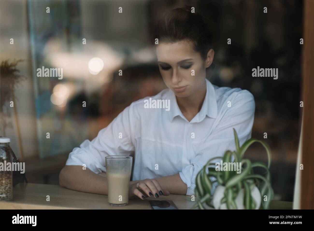 Belle jeune fille s'assoit dans un café près de la fenêtre derrière un verre avec une tasse de café et envoie un message sur le smartphone. Photo à travers verre avec Banque D'Images