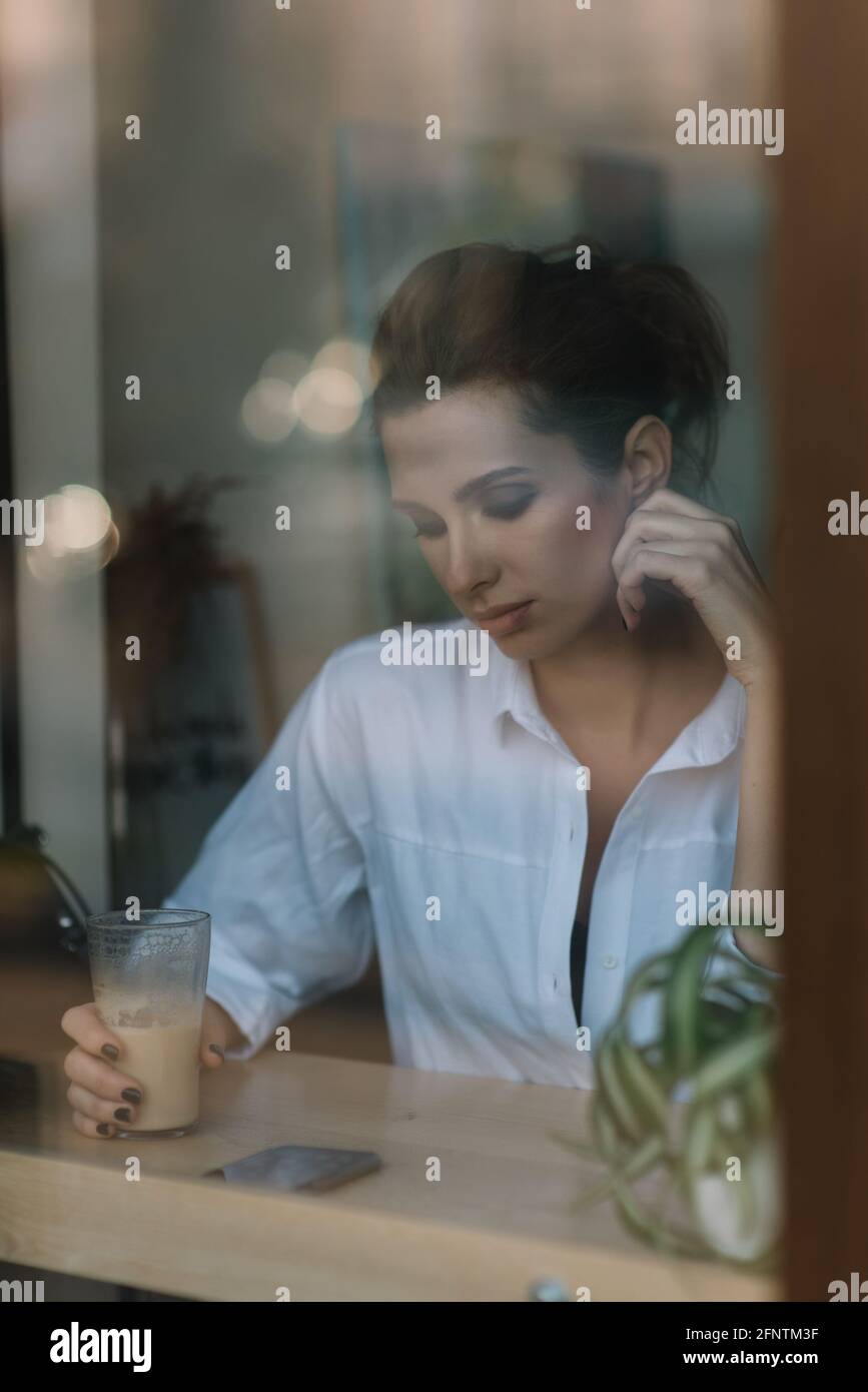 Belle jeune fille est assise dans un café derrière un verre avec une tasse de café et regarde dans un smartphone. Photo à travers le verre avec réflexion. Sel. Doux Banque D'Images