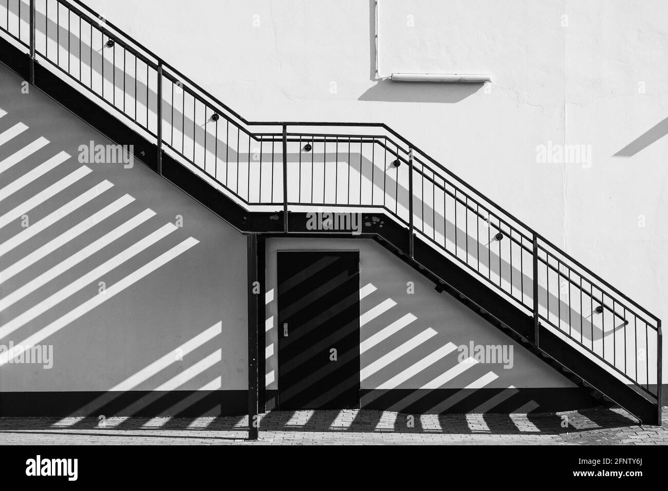 Un escalier extérieur s'étend en diagonale le long du mur de la maison et projette des ombres sur le mur et le sol pavé - en noir et blanc. Banque D'Images