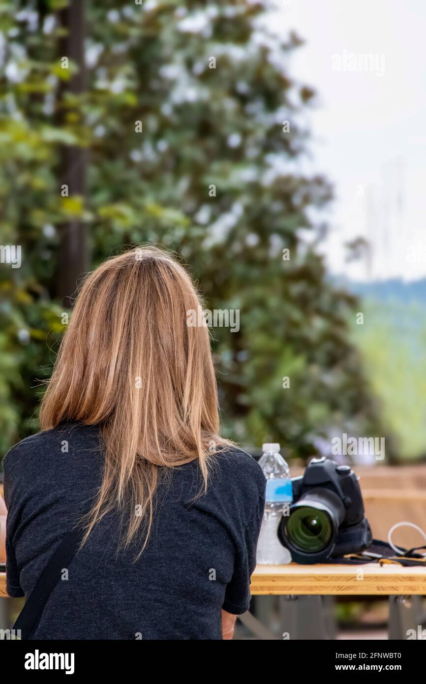 Arrière de la femme avec de beaux cheveux longs striés assis à table de pique-nique extérieure avec appareil photo et bouteille d'eau à côté elle avec un arbre bokeh dans backgrou Banque D'Images