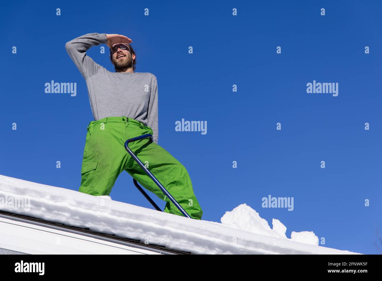 Portrait amusant et emblématique d'un jeune homme debout sur le toit d'une maison pendant un travail de déneigement, posé avec la main sur les sourcils Banque D'Images