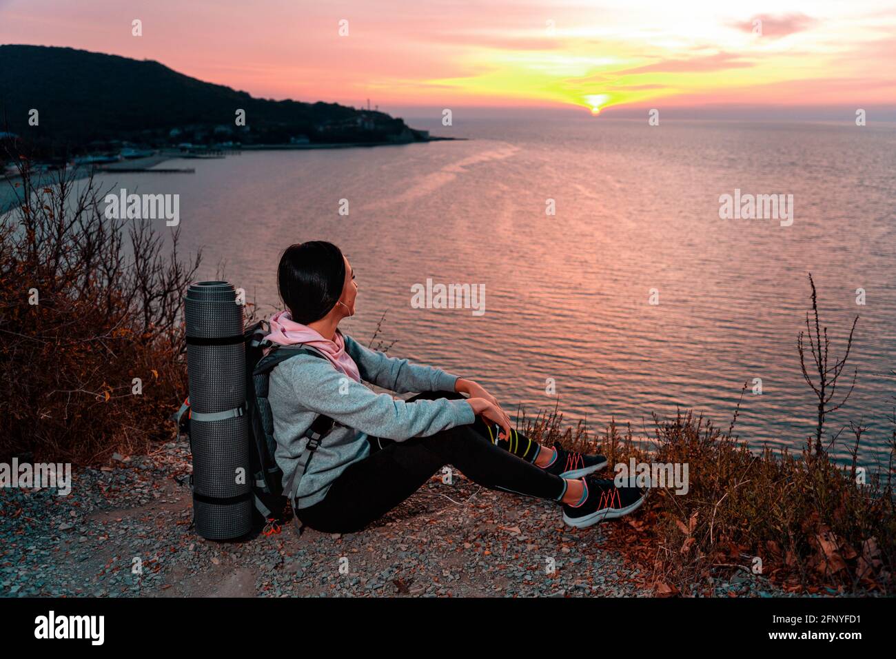Zeg opzij Wat snijden Une jeune femme avec un sac à dos sur son dos, assise au bord d'une falaise  et admirant le coucher du soleil. En arrière-plan, la mer, la montagne et  la sk Photo