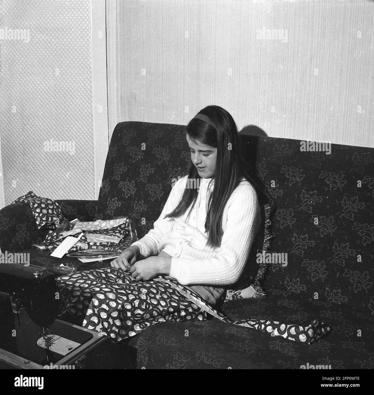 1960, historique, une adolescente assise sur un canapé en train de coudre à la main éventuellement sur des housses de coussins, avec une machine à coudre en face d'elle, Angleterre, Royaume-Uni. Banque D'Images