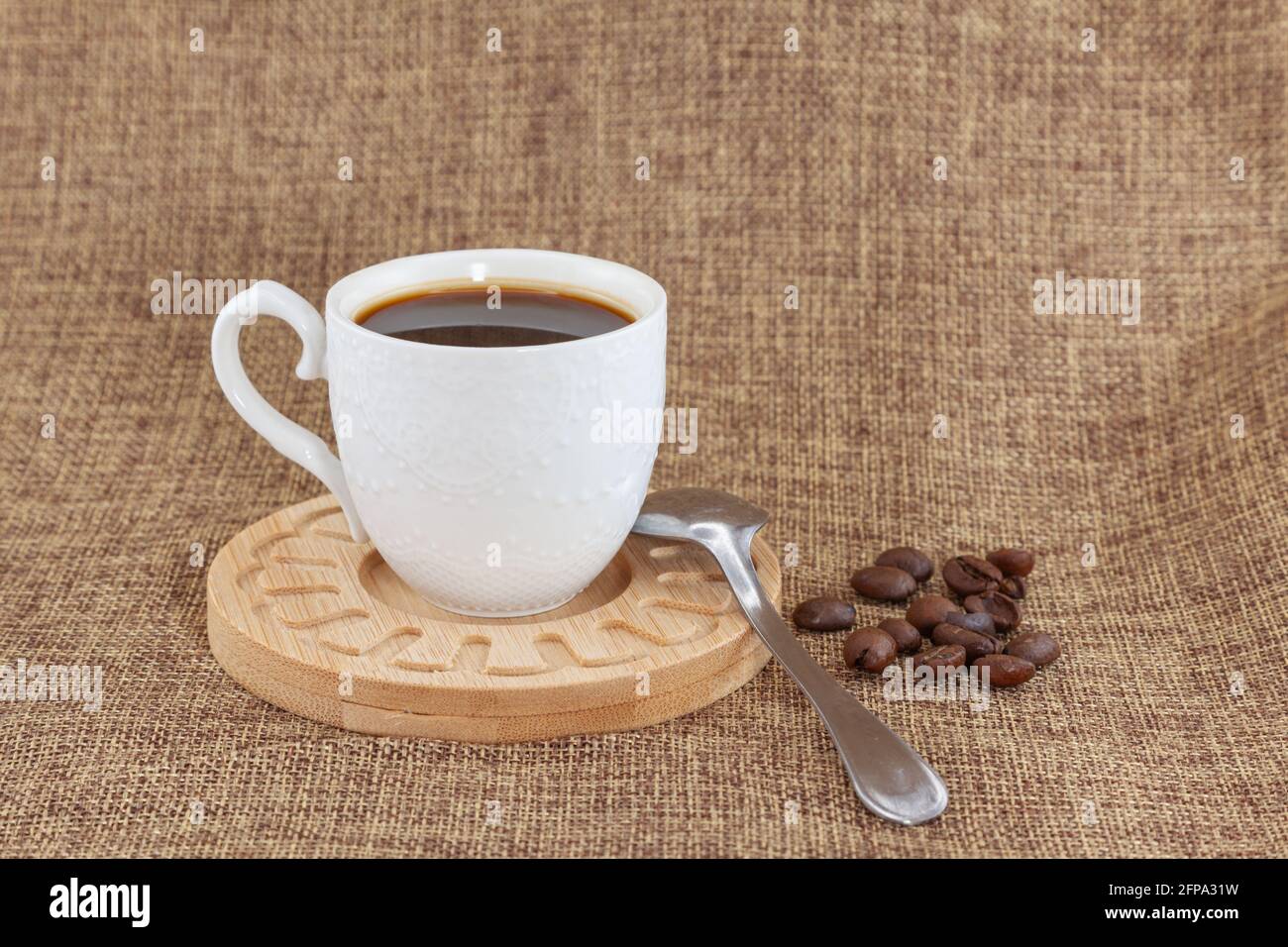 Sur une assiette en bois il y a une tasse blanche avec du café et une cuillère en métal. Ils sont placés sur un chiffon et il y a une poignée de grains de café sur le ri Banque D'Images