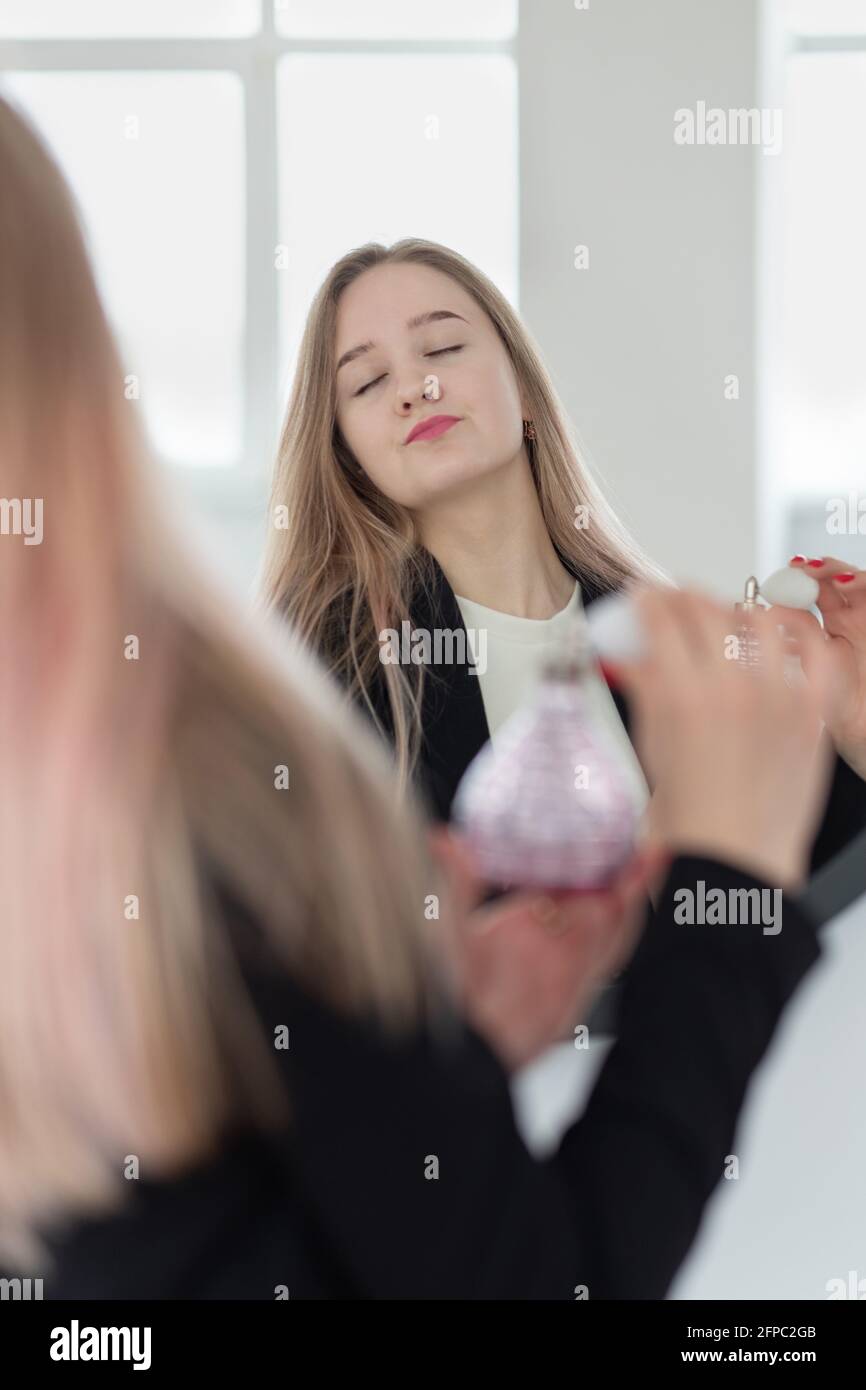 Jeune femme blonde caucasienne tient une bouteille de parfum ou d'eau de toilette et saupoudrer de poire en spray sur elle-même, regarder dans le miroir, fermer les yeux, le mode de vie. Banque D'Images