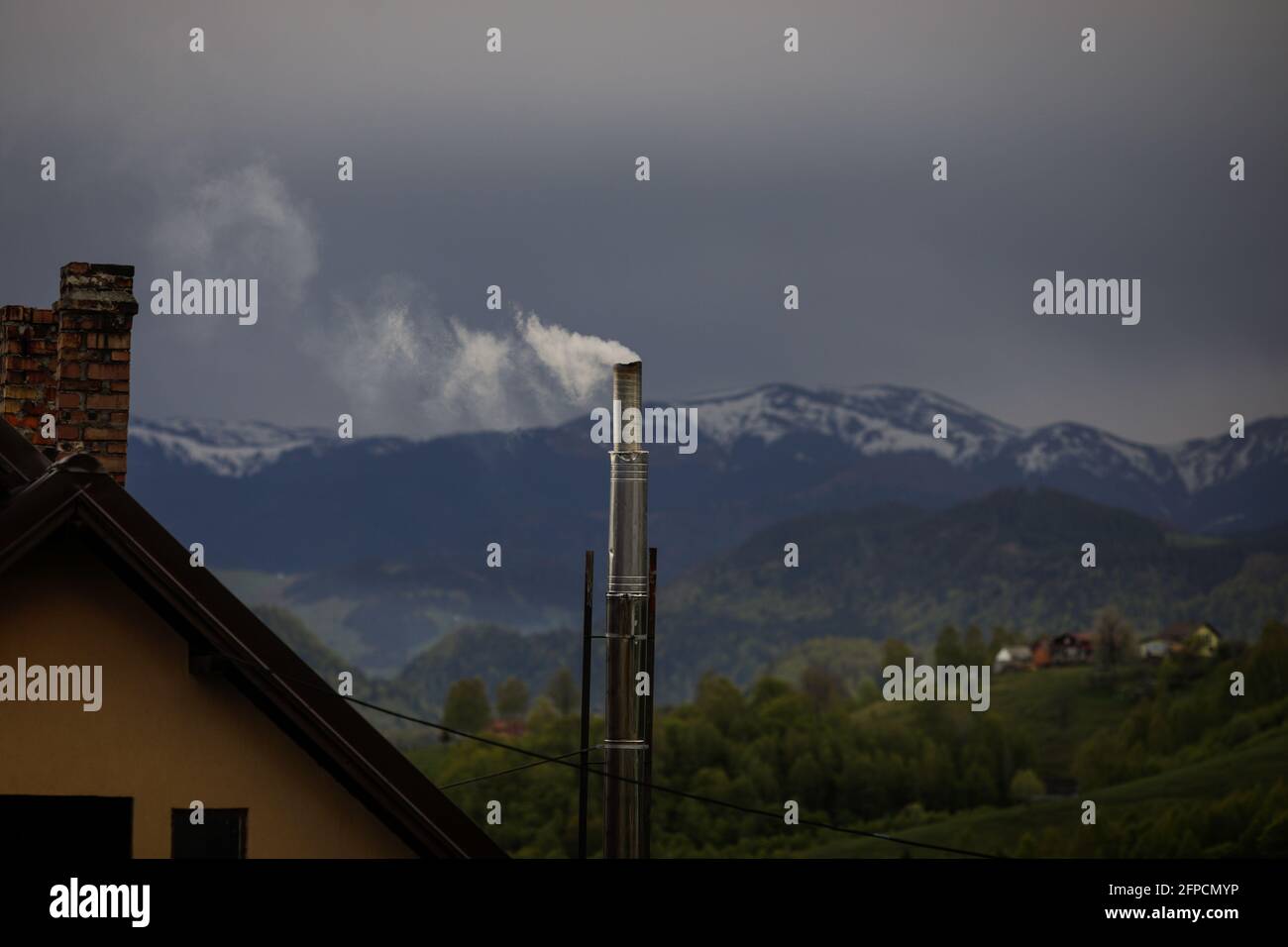 Fumée sortant d'une cheminée métallique avec les montagnes Bucegi en arrière-plan, en Roumanie rurale, au crépuscule du printemps. Banque D'Images