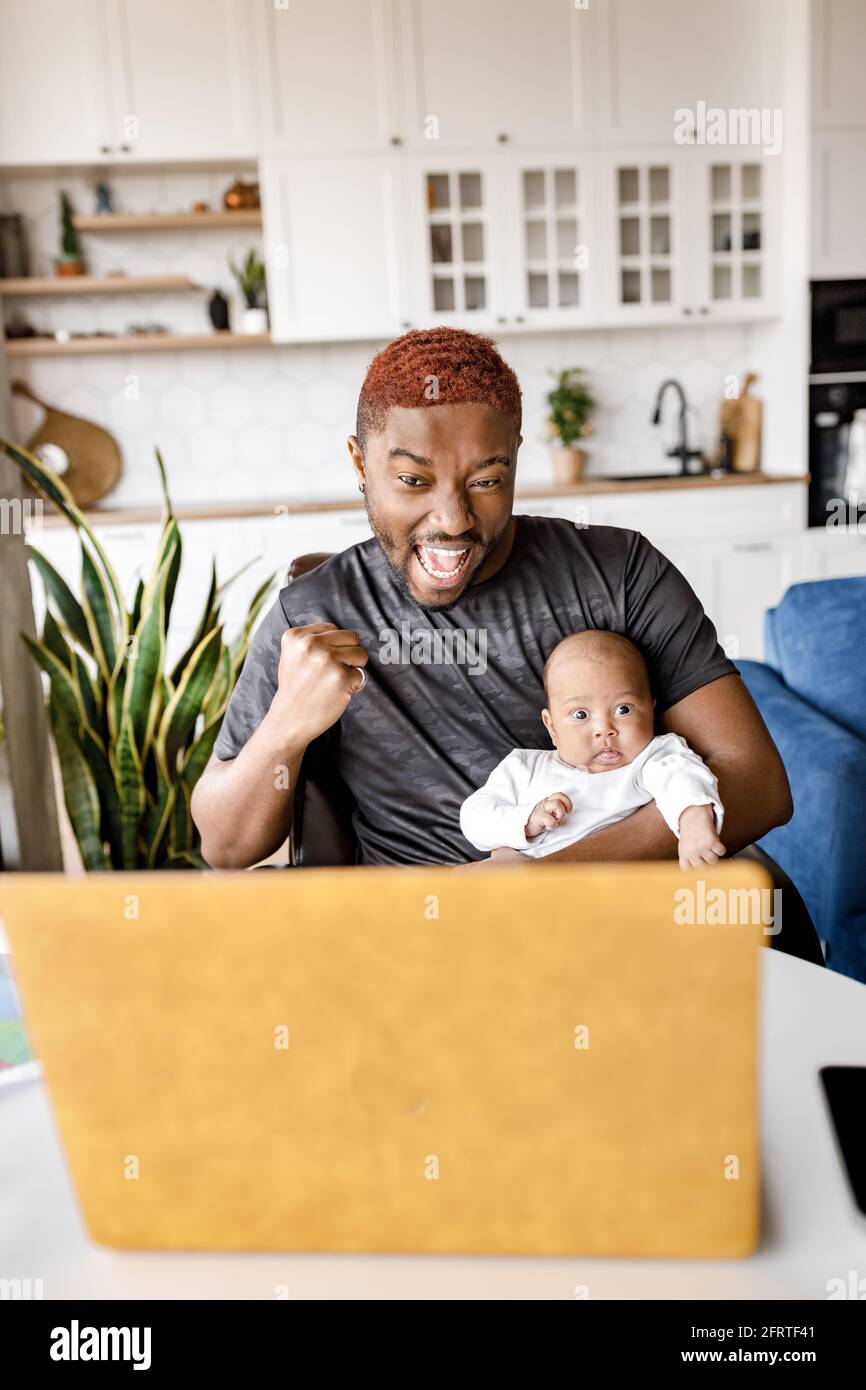 Un père afro-américain joyeux avec bébé son regardant l'écran d'un ordinateur portable, célébrant des nouvelles incroyables et réussies en ligne au bureau. Émotionnel Banque D'Images