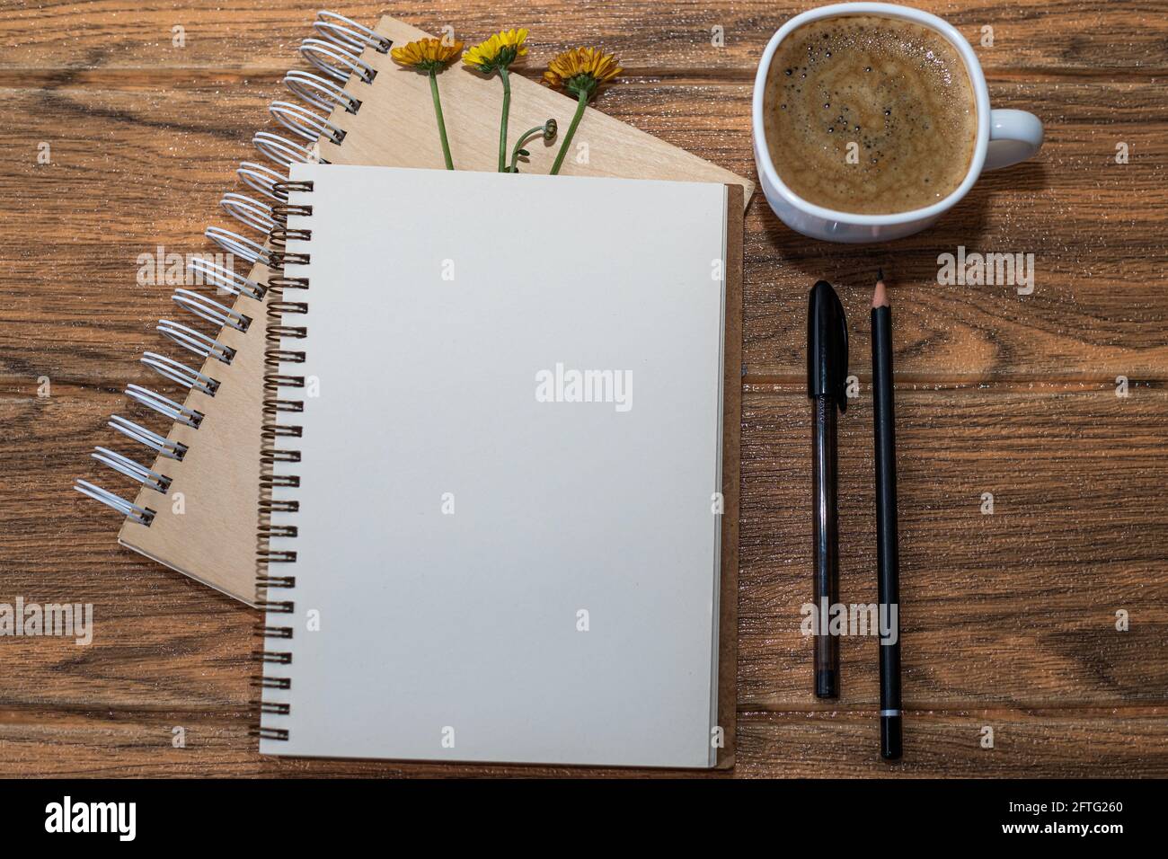 Carnets en spirale avec 3 fleurs. Une ouverture avec une feuille blanche et un stylo noir à côté. Il y a une autre tasse de café sur la table en bois. Banque D'Images