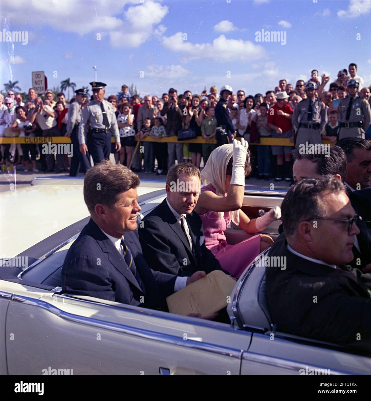 Le président John F. Kennedy et la première dame Jacqueline Kennedy se déplacent à l'arrière d'un cabriolet alors que leur cortège se rend au stade Orange Bowl à Miami, en Floride, pour une cérémonie de présentation du 2506e drapeau de la brigade d'invasion cubaine. Le président Kennedy tient sur ses genoux une grande enveloppe inscrite avec une note manuscrite, "Peech - Miami"; le maire de Miami, Robert King High, siège entre le président et Mme Kennedy (portant des gants blancs et un foulard enveloppé sous son menton). Sur le siège avant de la voiture (de gauche à droite) : agent du service secret de la Maison Blanche, Floyd Boring. Banque D'Images