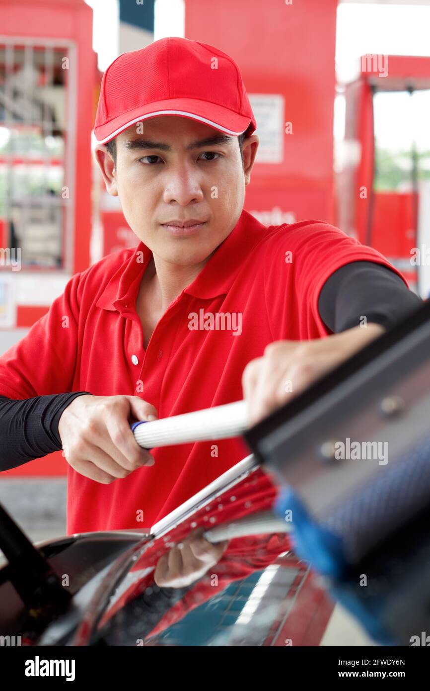 Ouvrier de station-service asiatique en uniforme rouge nettoyant le pare-brise de voiture avec raclette de fenêtre. Banque D'Images