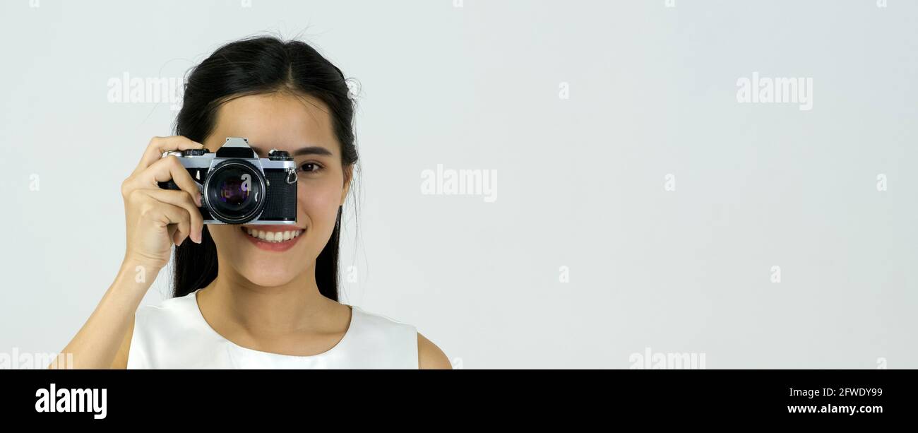 Jeune femme asiatique en robe blanche regardant à travers le viseur tout en filant avec l'appareil photo. Banque D'Images