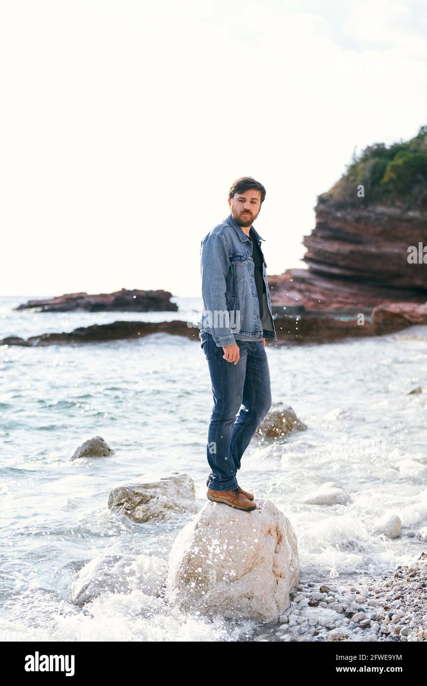 Homme pensif en Jean et une veste en denim se tient dessus une grande pierre et donne sur la plage de galets sur fond de montagnes rocheuses Banque D'Images
