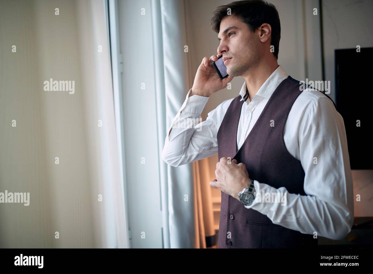 un jeune homme de race blanche, attentif, parle sur un téléphone portable près de la fenêtre et attache des boutons sur sa veste avec une autre main Banque D'Images
