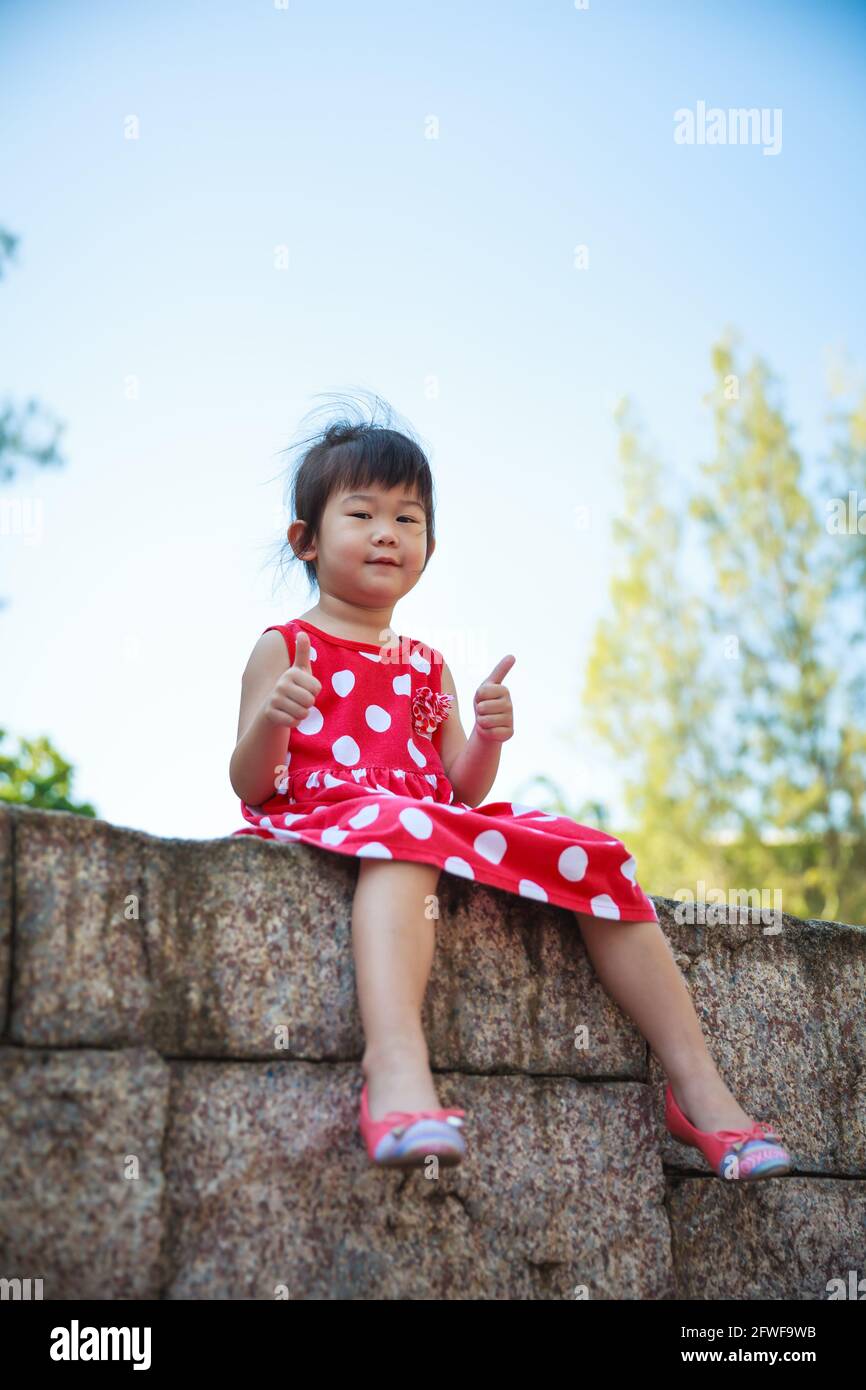 Bonne fille asiatique avec thump up et regardant l'appareil photo au parc. Enfant se détendant à l'extérieur dans la journée, voyage en vacances. Émotion humaine positive. Banque D'Images